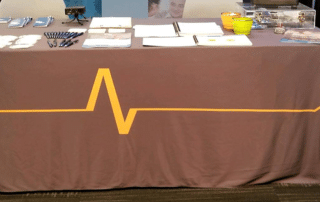 A booth table covered with a gray cloth featuring a yellow heartbeat line. On top of the table, there are various items, including brochures from Activated Insights, notebooks, pens, plastic cups, and other promotional materials. The background shows a glimpse of a partly visible banner.