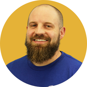 Headshot Template (2) A bearded man with a shaved head is smiling at the camera. He is wearing a blue shirt. The background is a solid mustard yellow in a circular frame.