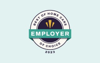 A circular badge with a light blue background reads "Best of Home Care Employer of Choice 2023". It features a dark blue center with three yellow bars and a green ribbon across the middle with the word "Employer".