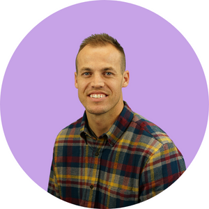 Jonalyn_Kelman (1) A man with short hair is smiling at the camera. He is wearing a colorful plaid shirt and is posed against a solid lavender circular background.