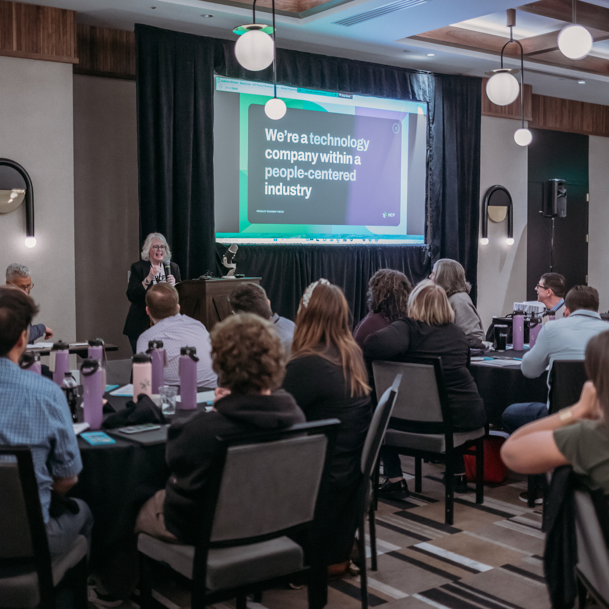A speaker is presenting to an audience in a conference room. On the screen behind the speaker, a slide reads, "We're a technology company within a people-centered industry." Attendees are seated at tables, listening attentively. The room has modern decor and lighting.