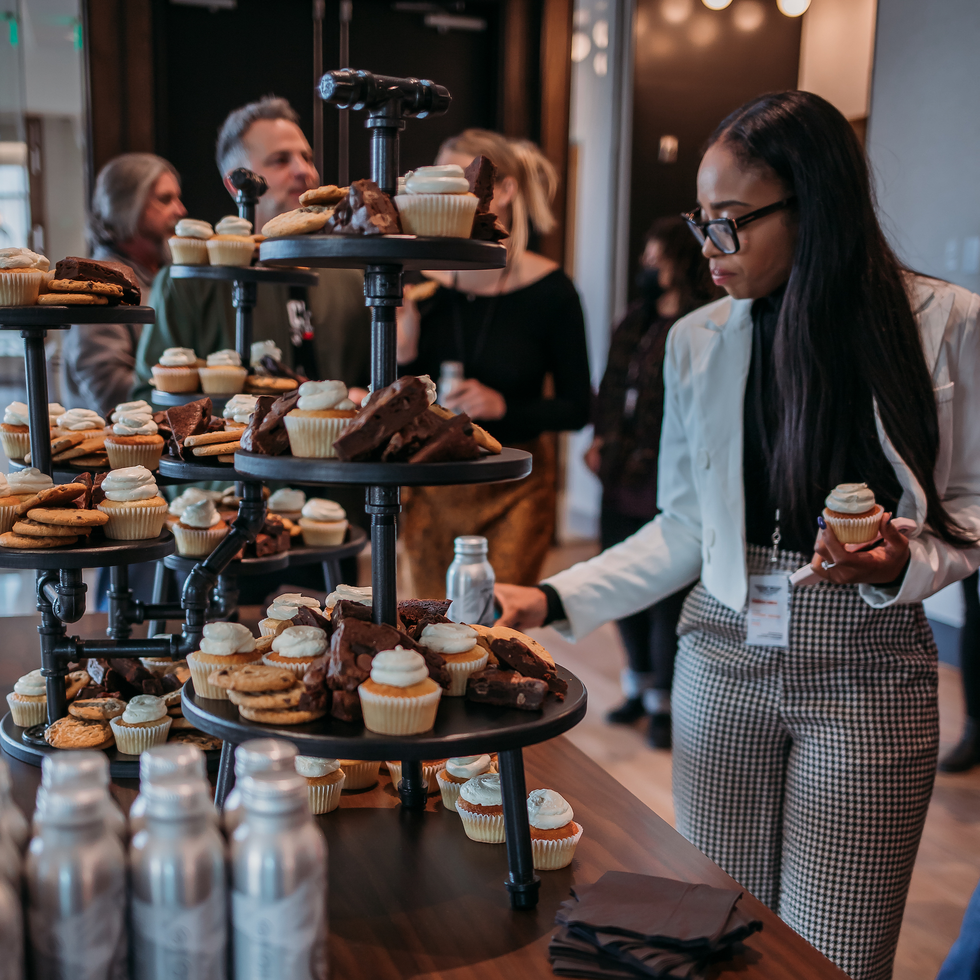 A woman in glasses and a light-colored blazer is holding a cupcake and a bottled drink while reaching for more baked goods from a multi-tiered dessert stand. Other people, casually dressed, are conversing in the background of a well-lit room, possibly at an event.