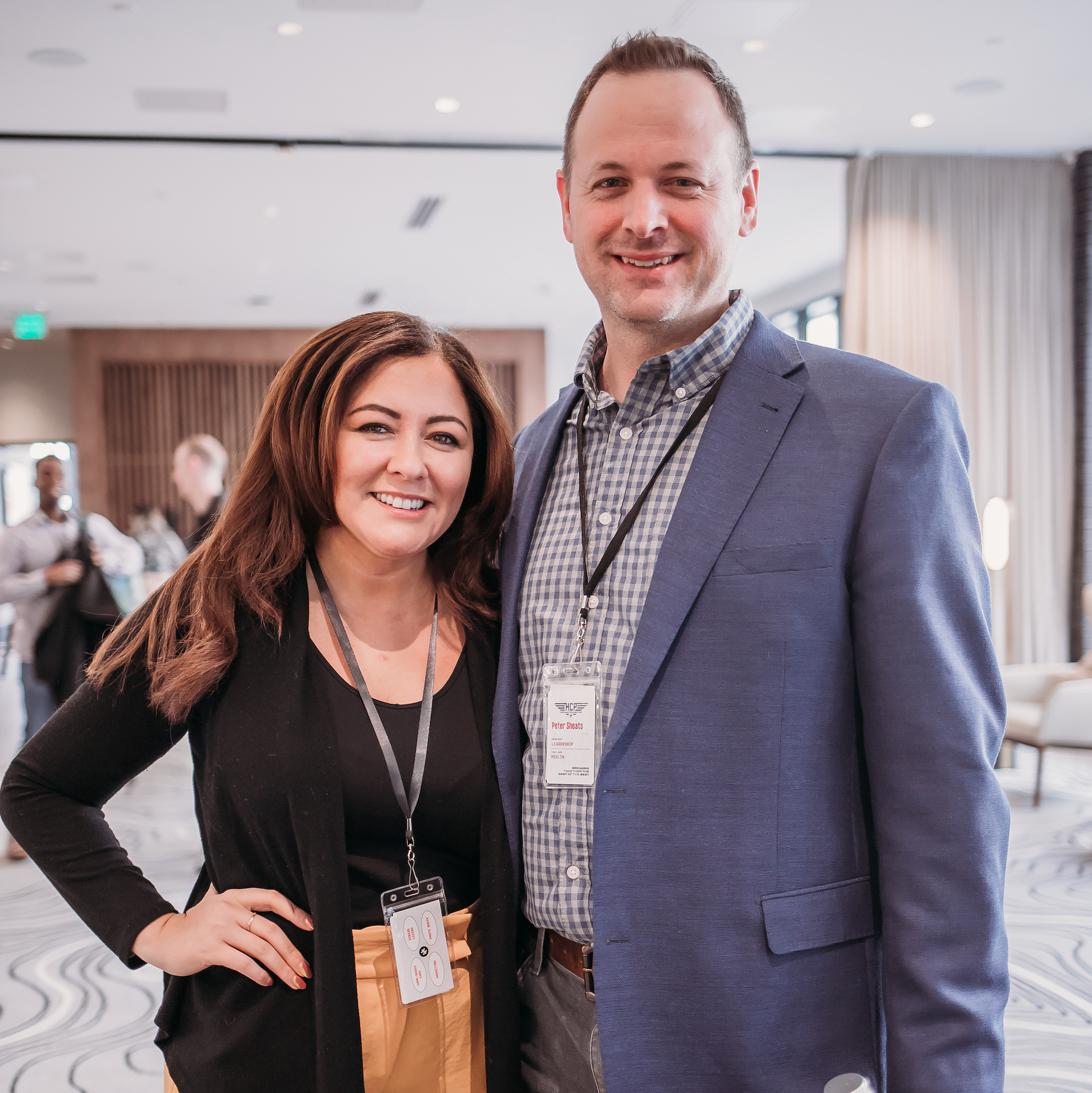 Two people are standing and smiling at the camera in a professional setting. The person on the left has long brown hair, is wearing a black top and yellow pants, and has a lanyard around their neck. The person on the right is dressed in a blue blazer with a checkered shirt.