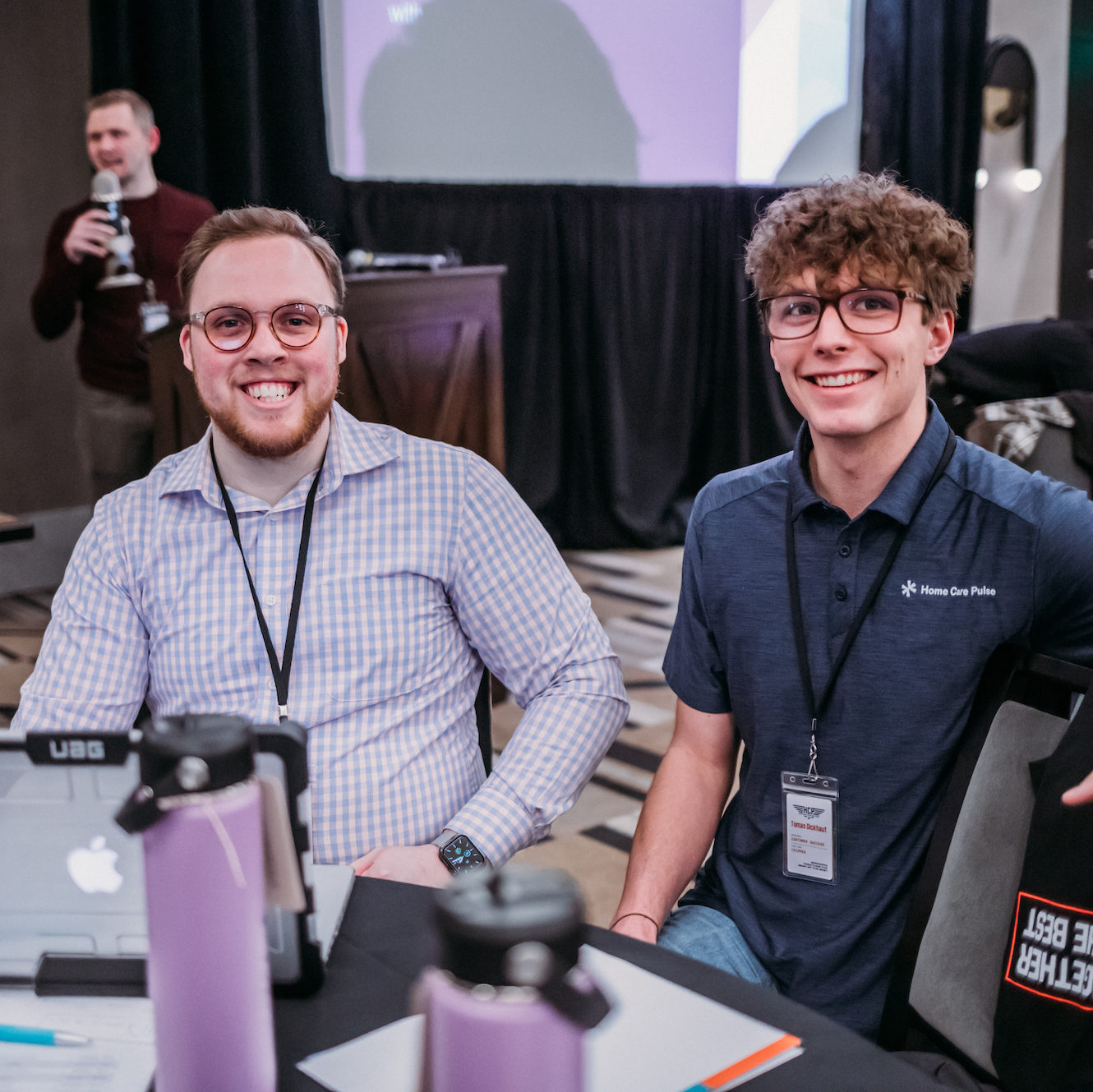 Two men are seated at a table during a conference, smiling towards the camera. The man on the left wears glasses and a checkered shirt, while the man on the right wears glasses and a polo shirt. Conference materials and water bottles are on the table. Another person stands in the background.