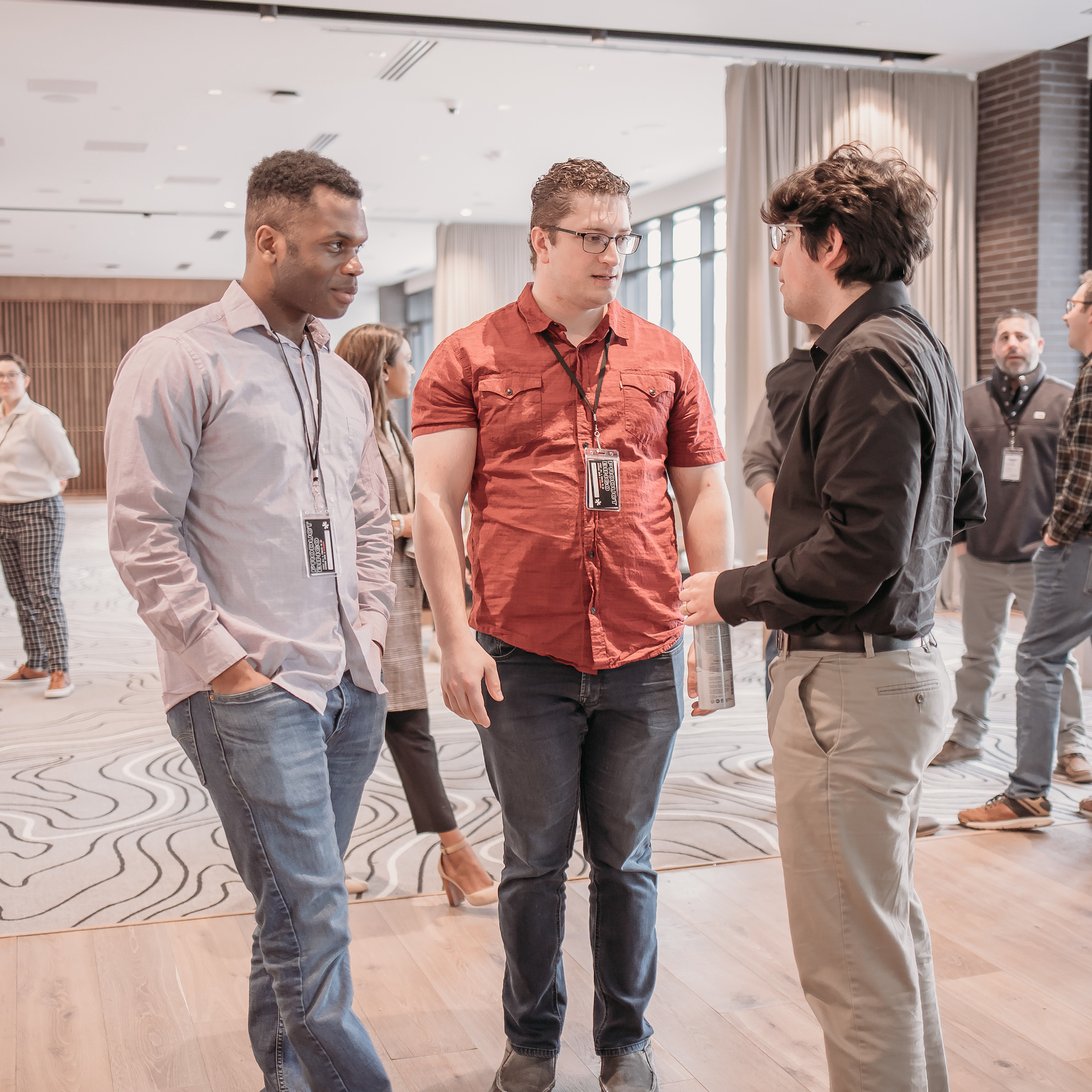 Three men are having a conversation at a networking event in a bright, modern room. They are wearing name badges, business casual attire, and are standing on a patterned carpet. Other people can be seen in the background, also engaged in discussions.