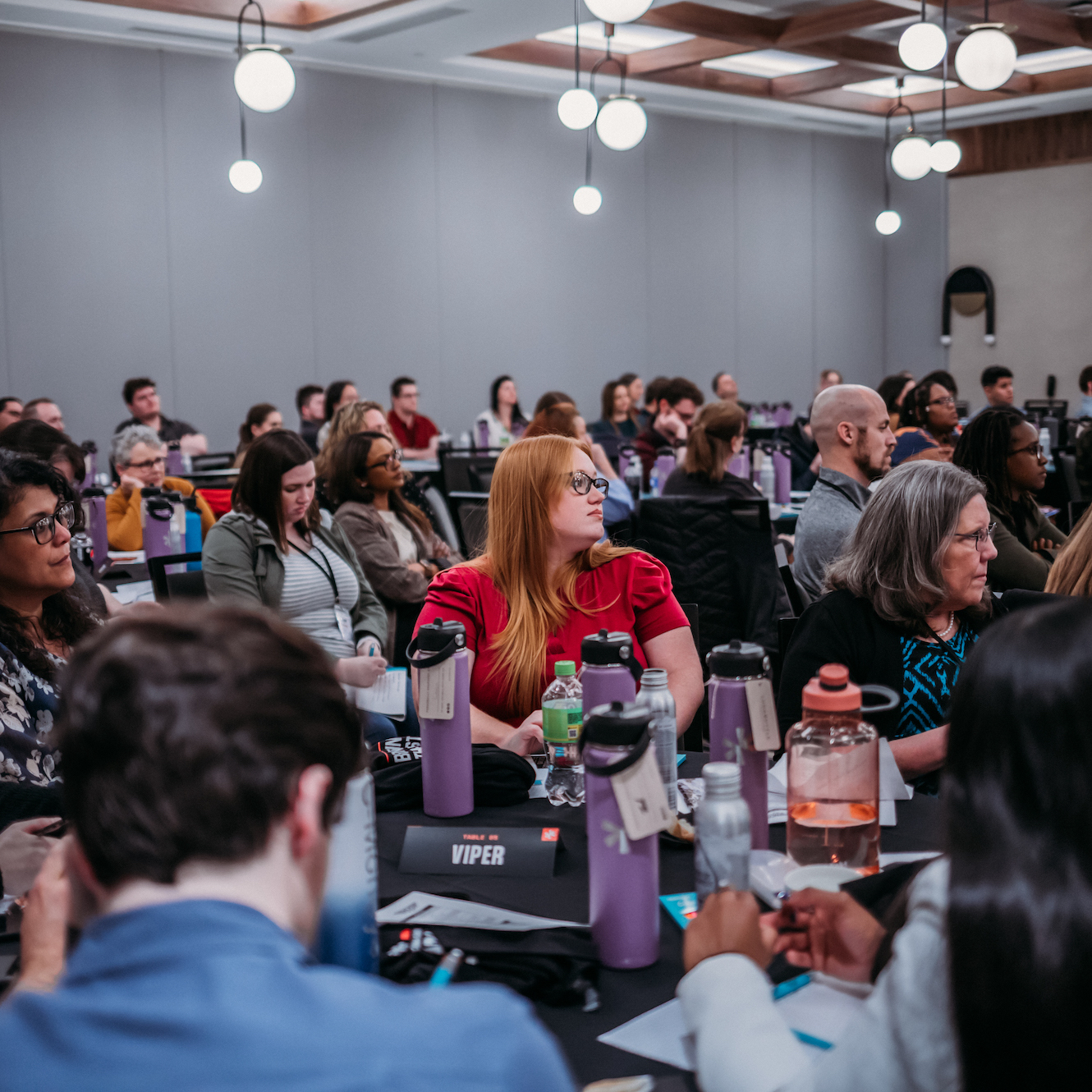 A diverse group of people sits attentively in a conference room. Some take notes while others listen to a speaker out of the frame. The room has modern light fixtures, and attendees have water bottles and notebooks on the tables in front of them.