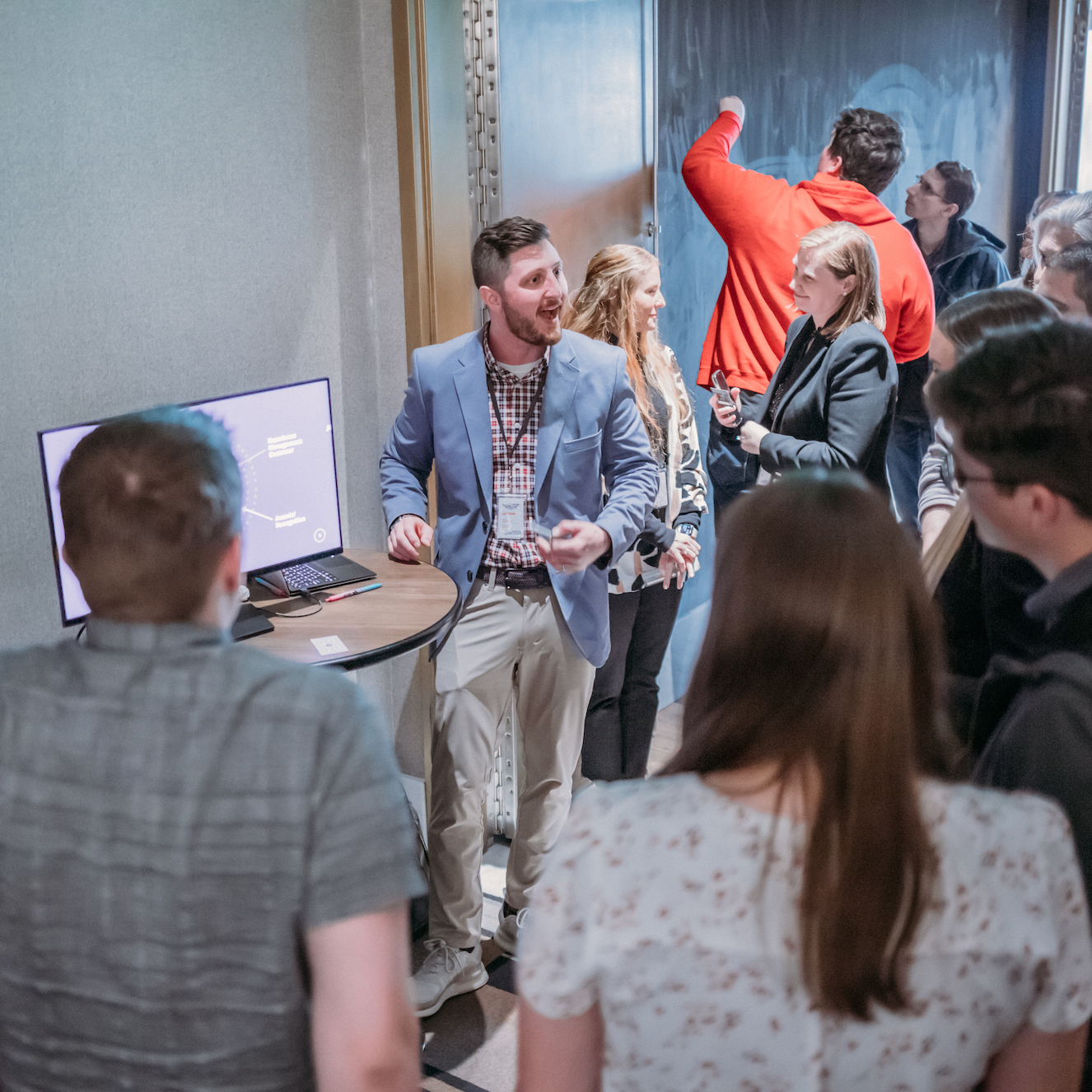 A group of people in a room, with a man in a suit and a name tag speaking near a table with a computer monitor. Some individuals are interacting in the background. The setting appears to be a professional or networking event.