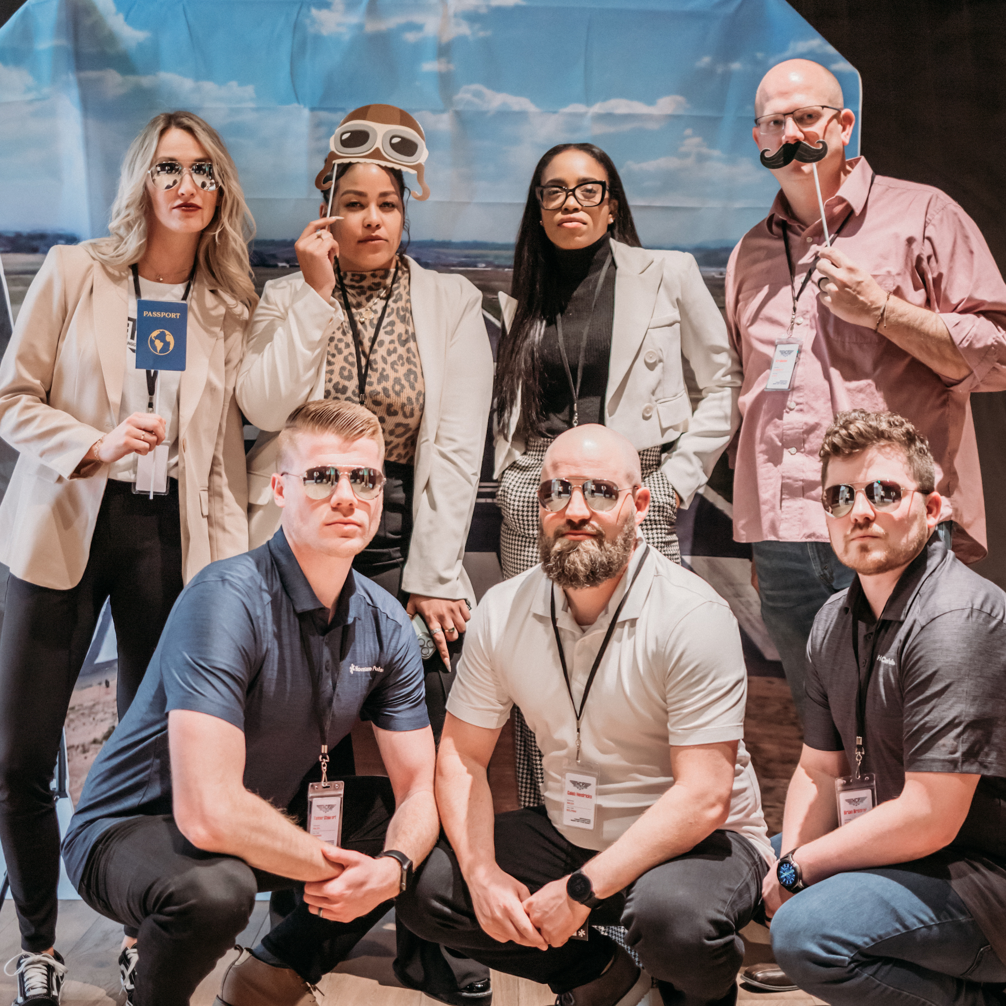 A group of six people posing together, dressed in casual and semi-formal attire. Four are kneeling in front, while two stand behind, one holding a prop passport and another wearing aviator goggles. All are wearing reflective sunglasses, giving a fun, unified look.