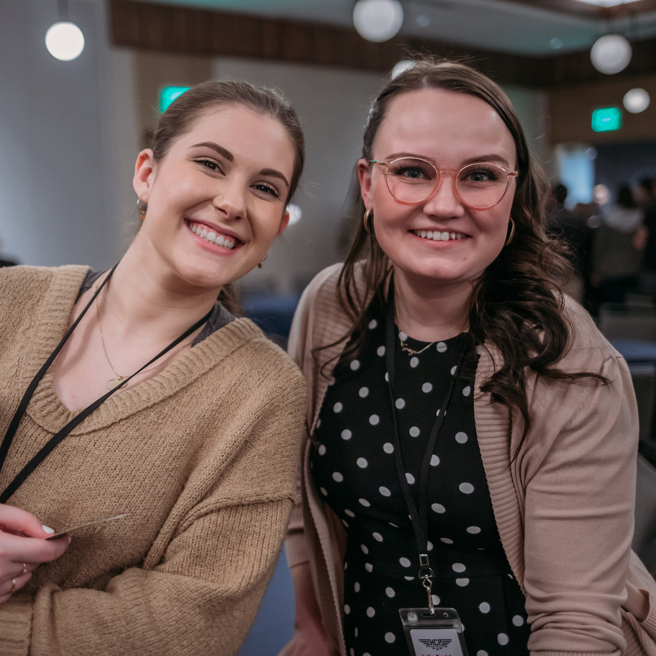 Two smiling women are pictured indoors. The woman on the left is wearing a beige sweater and a black lanyard, while the woman on the right has glasses, a polka-dot dress, and a beige cardigan, also with a lanyard. Both are looking at the camera.