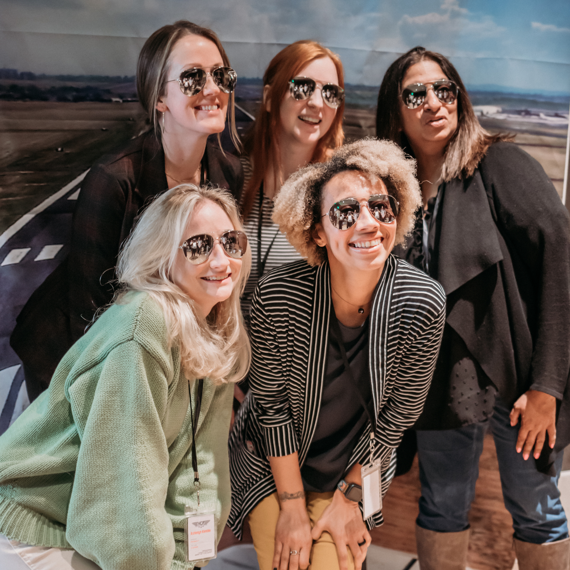A group of five women, all wearing sunglasses, smiles and poses together in front of a backdrop depicting an airport runway. They are dressed casually and appear to be enjoying their time together.