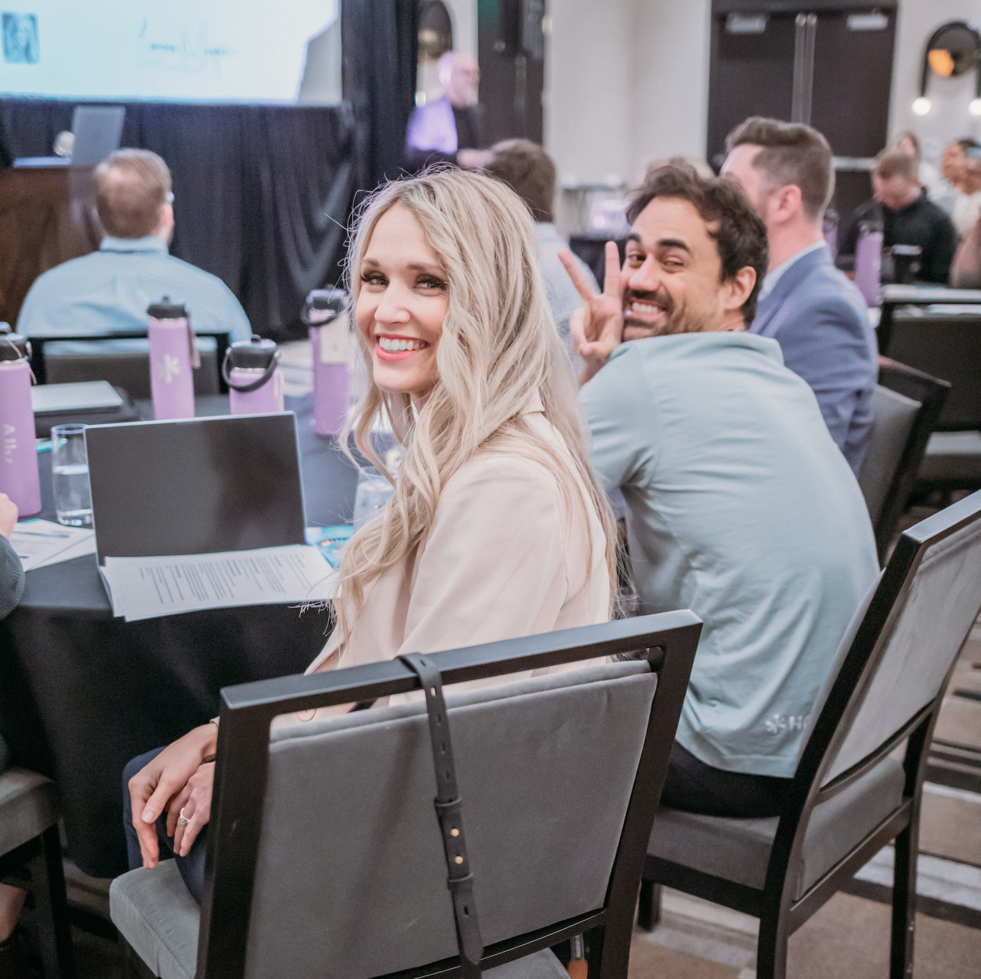 A woman with long blonde hair and a man smiling and gesturing a peace sign are seated at a conference table in a meeting room. Attendees in business attire face a speaker at the front. Purple water bottles and documents are on the table.