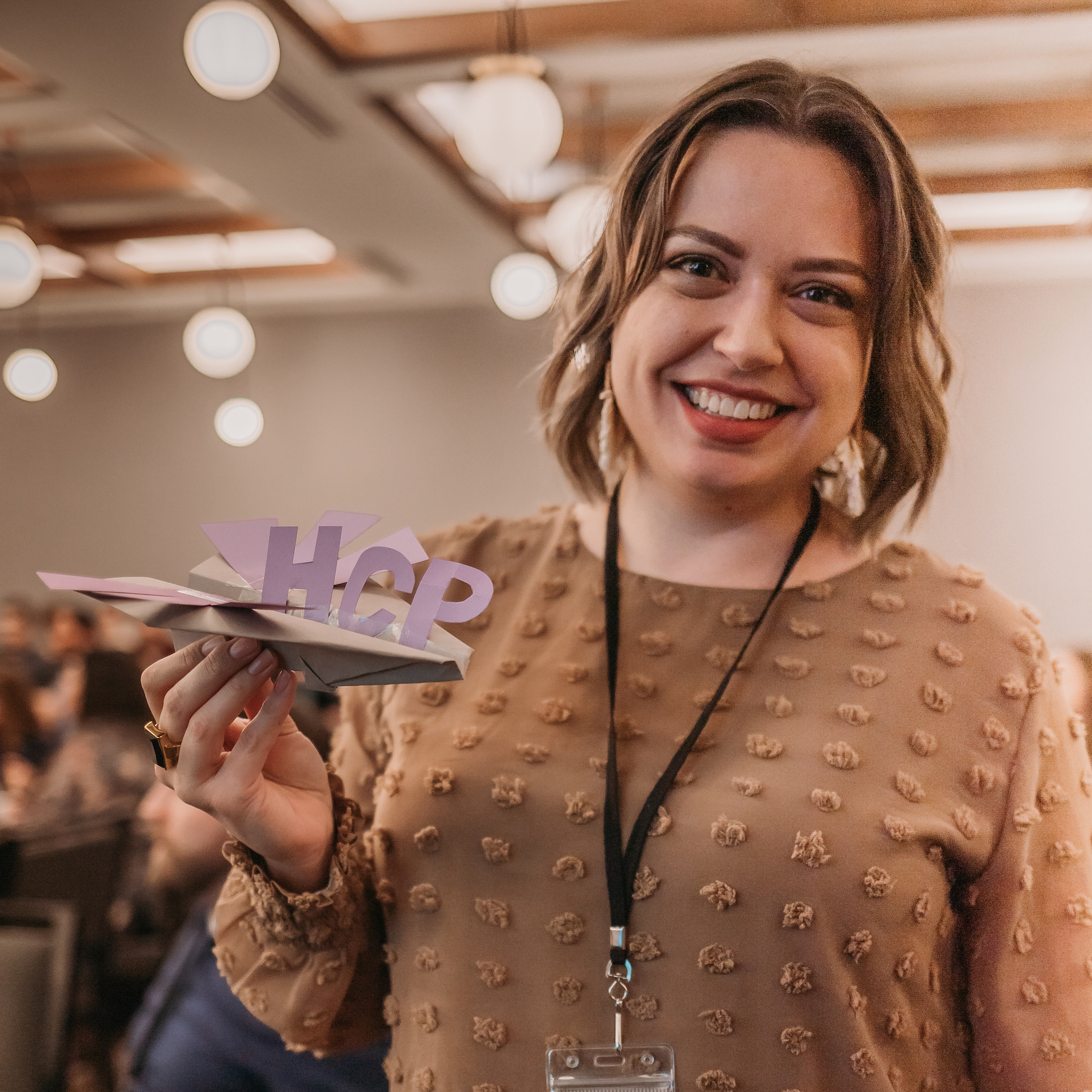 A woman with shoulder-length hair is smiling and holding a paper airplane with "HCP" written on it. She is wearing a brown, textured blouse and a lanyard. The room behind her is out of focus, with round ceiling lights.