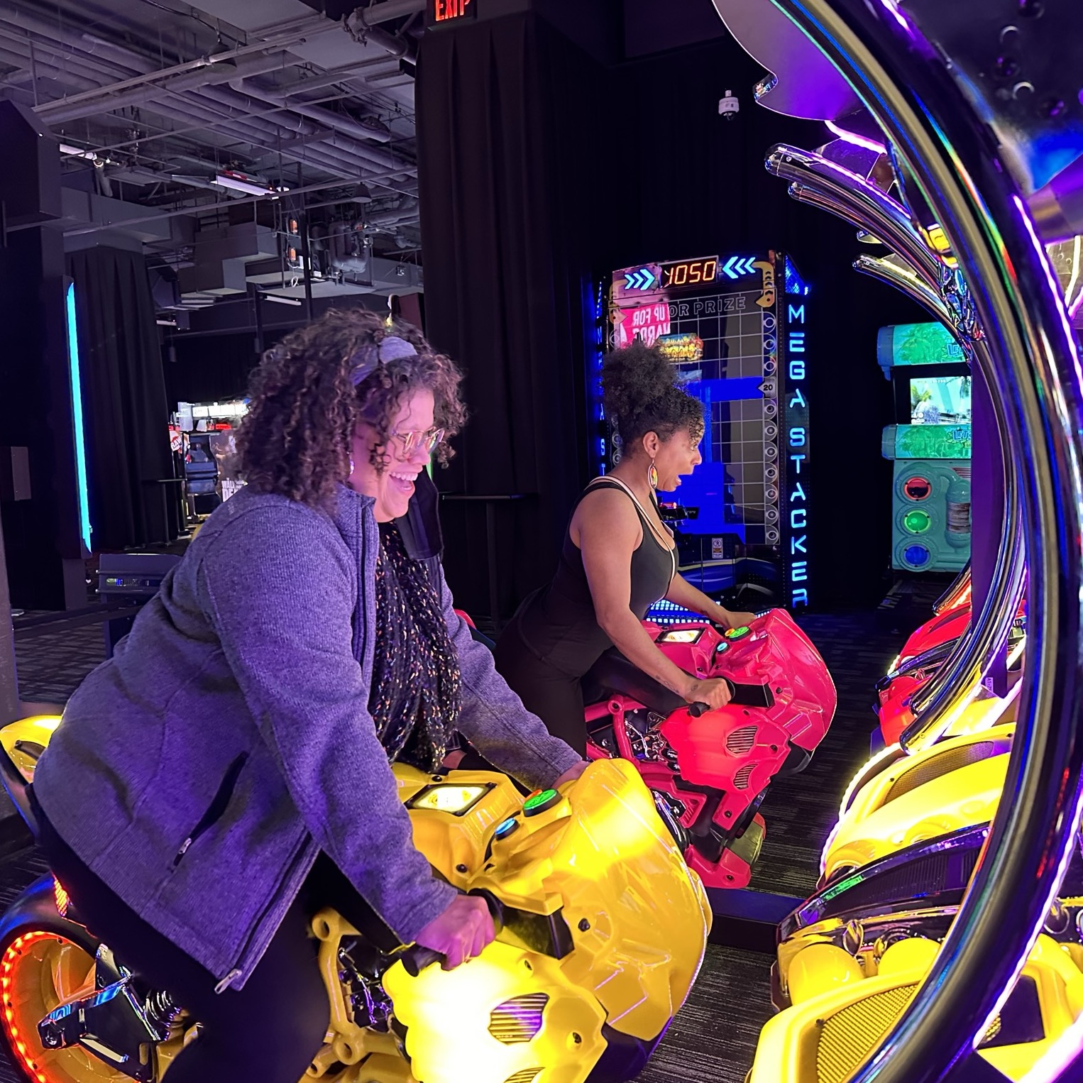 Two people are playing a motorcycle racing arcade game. They are seated on yellow and red motorbike consoles with colorful lights. The background features other arcade machines and games, creating a lively and energetic atmosphere.