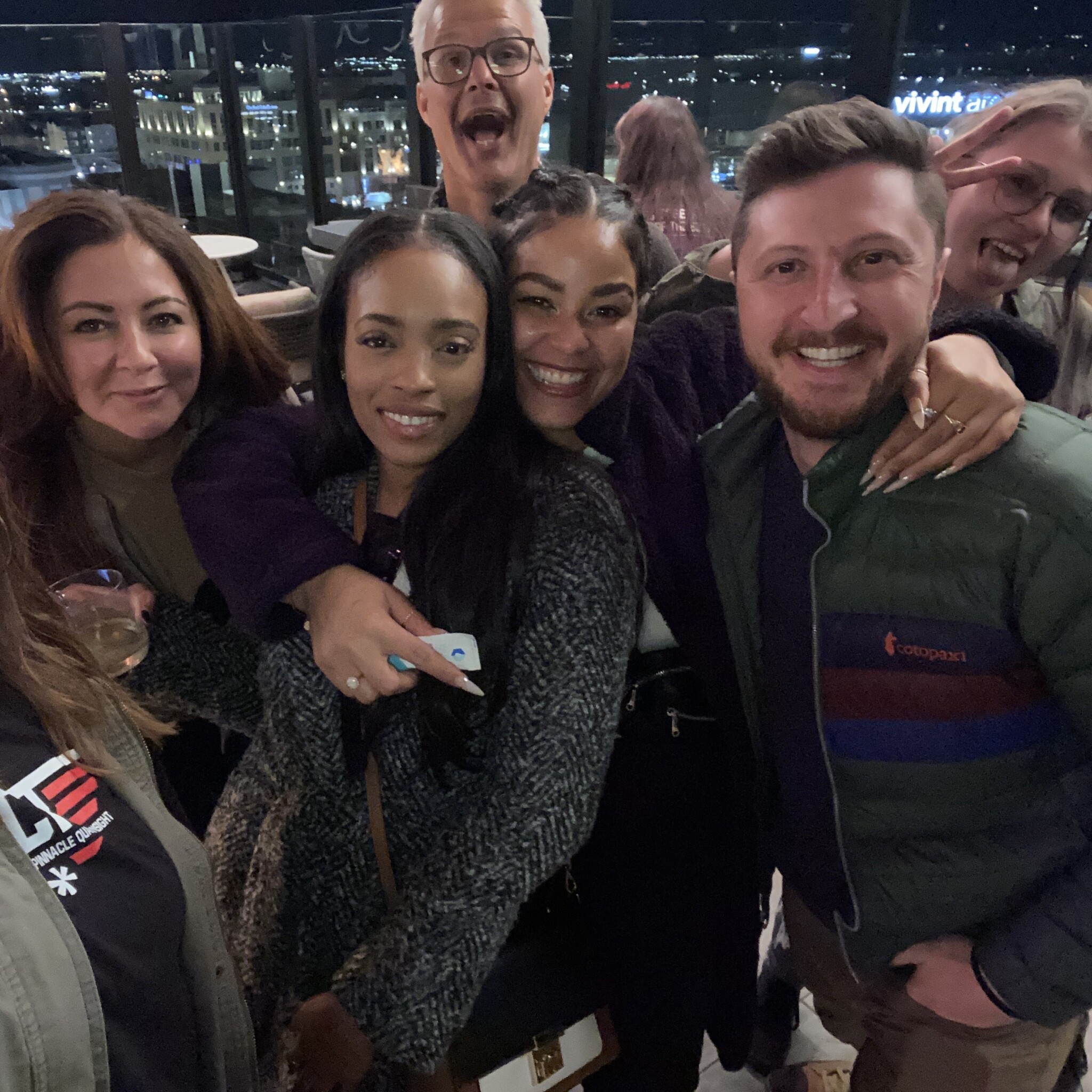 A group of six people are standing closely together and smiling for a photo. One person in the background is making a funny face with their mouth wide open. The background shows a nighttime cityscape with lights.
