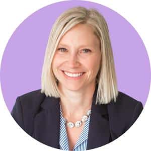 Sarah Dirks A woman with shoulder-length blonde hair is smiling. She is wearing a dark blazer over a blue and white striped shirt, accessorized with a silver necklace. The background is a light purple circle.