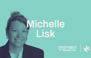 A professional headshot of a person with short hair pulled back. They are smiling and wearing a white shirt and dark blazer. The background is teal, and the text reads "Michelle Lisk." Below is the SYNERGY HomeCare logo and an asterisk symbol.