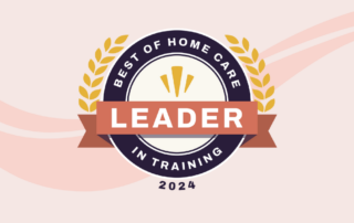 A circular badge logo with gold laurel leaves on either side. It reads "Best of Home Care" at the top, with "Leader" in the center on a red ribbon, and "In Training" below. The year 2024 is at the bottom. The background has soft pink and peach wavy lines.