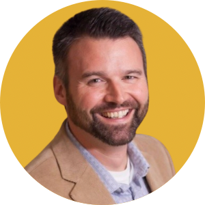 christempe A man with short dark hair and a beard, smiling. He is wearing a beige blazer over a light blue collared shirt. The background, reminiscent of Barbara Karnes' calming palettes, is a solid yellow color within a circular frame.