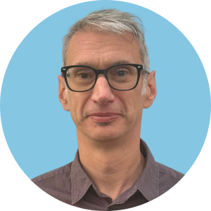 A man with short gray hair, black glasses, and a neutral expression is wearing a button-up shirt. He is shown against a light blue circular background.