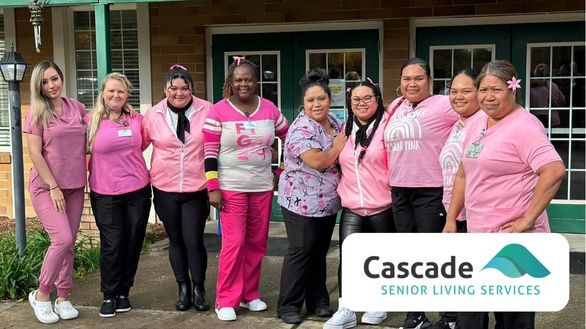 A group of nine women dressed in pink stand smiling outside a building. They appear to be staff members. A logo in the corner reads "Cascade Senior Living Services.