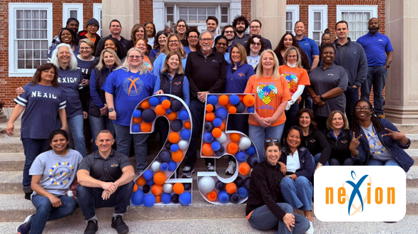 A diverse group of people poses outside a building, smiling around a decorated "25" made of balloons. The Nexion logo appears in the bottom right corner, marking a 25th anniversary celebration.