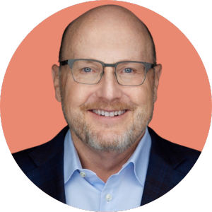 Smiling bald man with glasses, light beard, and mustache, wearing a blue collared shirt and dark blazer, against a solid peach-colored circular background.