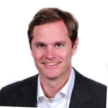 Scott Asher A man with short brown hair, wearing a dark pinstripe suit jacket and a white collared shirt, smiles at the camera against a plain white background.
